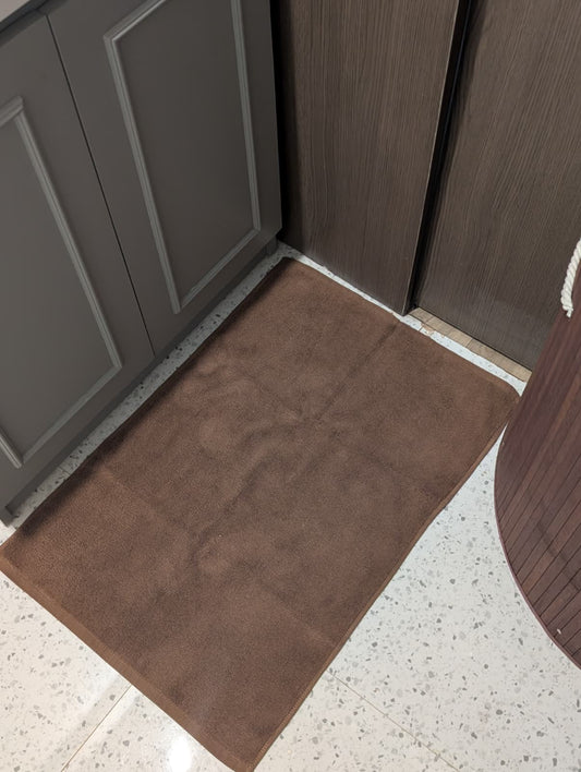 A rectangular brown bath mat or small rug placed on a speckled white floor between gray and dark wood cabinets.