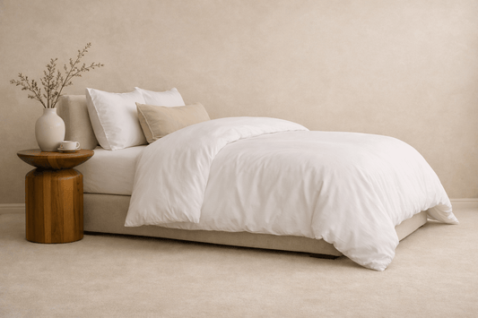 A neatly made bed with white bedding and two beige pillows, styled in a minimalist room with a small wooden side table.