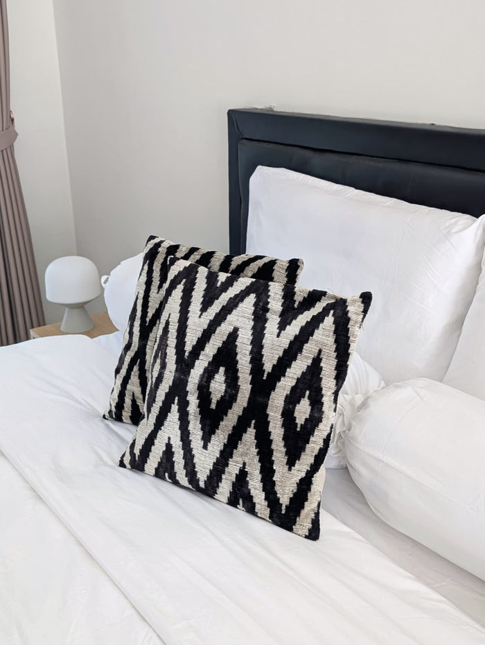 2 black and white decorative pillows on a white bedding set underneath and a small lamp 