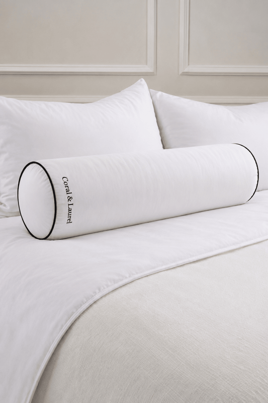 A white bolster pillow with black piping and embroidery on it, resting on a bed with white bedding and pillows.