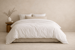 A neatly made bed with white bedding and several pillows, styled in a minimalist bedroom with a small wooden side table.