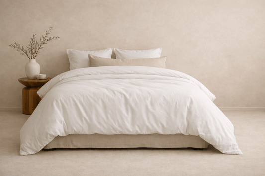 A neatly made bed with white bedding and several pillows, styled in a minimalist bedroom with a small wooden side table.