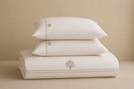 Three stacked white pillows and a folded white sheet or pillowcase set on a beige surface against a beige background.