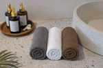 Three rolled bath towels in gray, white, and brown are arranged on a speckled stone countertop next to a large vessel sink.