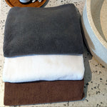 Three cotton towels: a dark charcoal gray towel on top, a white towel in the middle, and a brown towel on the bottom.