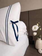 White bedding set with two pillows, one featuring two vertical navy blue stripes and a matching navy ribbon bow, on a bed.